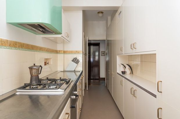 A narrow galley kitchen with cream cabinetry, long grey benchtops, a four-burner gas cooktop, stainless-steel rangehood, tiled walls, and open shelving. A hallway is visible beyond the kitchen, with a knife block and small appliances on the counters.