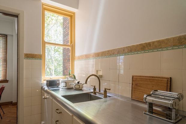 A bright traditional kitchen viewed along a long stainless-steel benchtop, with a deep double-bowl sink, curved gooseneck tap, light cabinetry, tiled splashback, timber-framed window, and countertop appliances. The cream wall tiles feature a decorative green border and textured horizontal band, while warm timber trims add character.