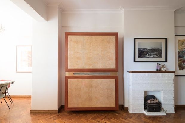 A bright traditional living room with a white-painted brick fireplace, timber mantel, framed artwork, parquet timber flooring, and a large built-in timber cabinet with textured panel doors. A glimpse of an adjoining dining area is visible to the left.
