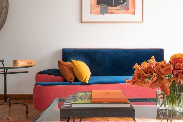 A colourful mid-century-inspired living room vignette featuring a deep blue and coral-red upholstered sofa with rust and mustard cushions, a glass coffee table with stacked books, a side table, artwork, and a vase of orange flowers. The room has white walls and warm timber flooring, with a bold, playful palette and strong contrast.