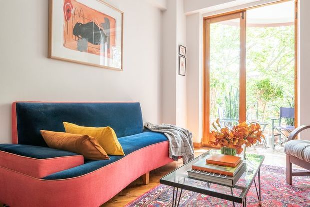 A bright, warmly styled living room featuring a bold coral-and-deep-blue upholstered sofa, colourful cushions, a glass-topped coffee table, patterned area rug, framed abstract artwork, and a large timber-framed glazed door opening to leafy greenery. A chair and small side tables are visible near the glazing.