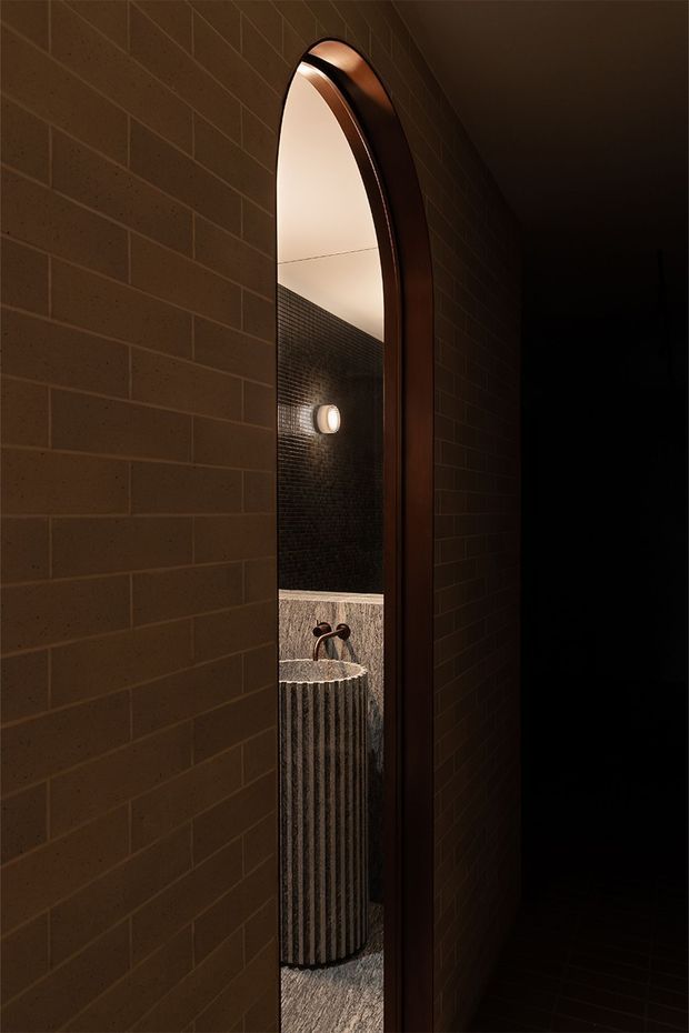 A narrow, moody interior passage with dark brown brick walls and a tall arched mirror in a dark timber or bronze-toned frame. The reflection reveals a compact bathroom vanity with a fluted stone pedestal basin, wall-mounted dark metal tapware, small dark mosaic tiles, and warm architectural lighting.