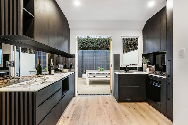 A contemporary galley-style kitchen with dark timber cabinetry, a pale stone island benchtop, integrated appliances, and light timber flooring. The kitchen opens through glazed doors to a small outdoor seating area, with white walls and recessed ceiling lighting creating a bright, refined contrast.