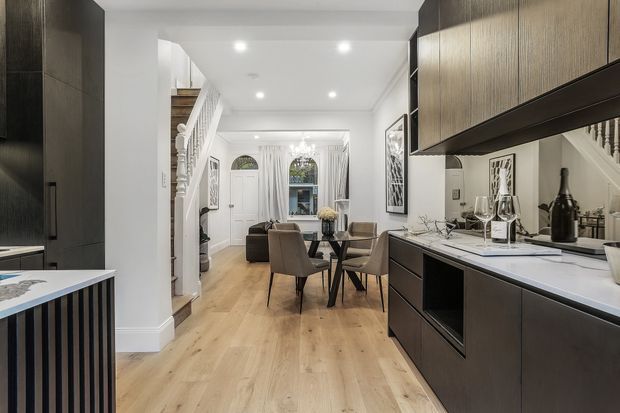Open-plan kitchen and dining area leading into a living room in a refined townhouse interior. The space features dark wood-toned kitchen cabinetry with a light stone benchtop, pale timber flooring, white walls and ceiling, a formal dining table with upholstered chairs, and a traditional stair and entry zone. Recessed ceiling lights and a central pendant provide illumination, while dark joinery creates contrast against the bright neutral interior.