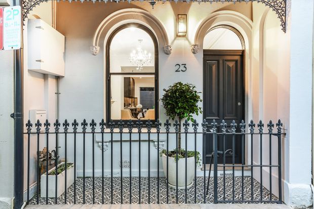 Street-facing entry porch of a narrow townhouse with an ornate rendered facade, arched openings, a black paneled front door, decorative black metal railings, patterned encaustic-style floor tiles, potted plants, and a glimpse into the interior through the arched window. The composition has a refined heritage character with warm neutral finishes and strong black detailing.