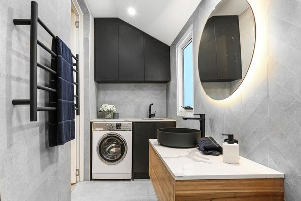 Compact contemporary bathroom with integrated laundry facilities. The room features large light-grey tiled walls and floor, matte black cabinetry, a black round vessel basin on a white stone-look benchtop, timber vanity drawers, a round mirror, black tapware, and a wall-mounted black heated towel rail.