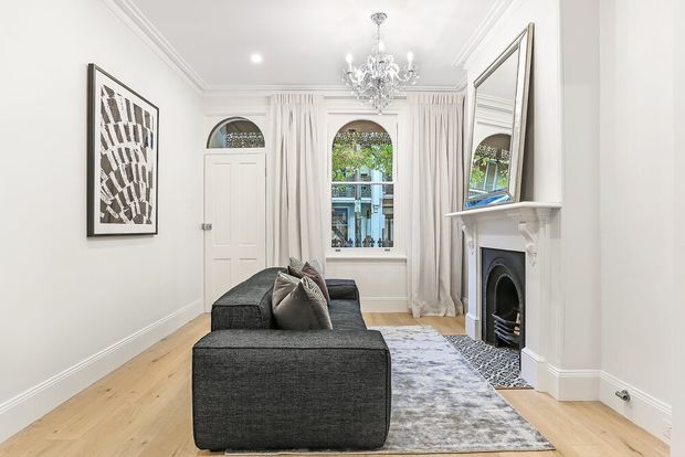 Bright, refined living room with a compact dark charcoal sectional sofa, pale timber flooring, a muted patterned area rug, ornate white fireplace mantel, large overmantel mirror, tall draped windows, decorative ceiling mouldings and a crystal chandelier. Arched window and door detailing add a traditional character, while the restrained furnishings give the room a contemporary feel.