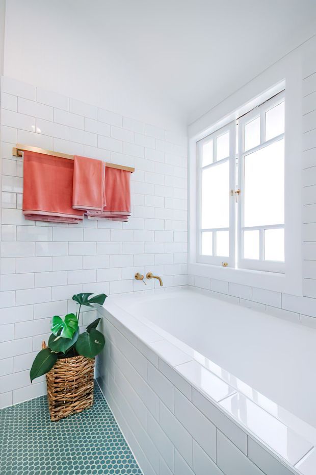 Bright, compact bathroom with white subway-tiled walls and bathtub surround, a white-framed window, brass wall-mounted bath tapware, and a small green mosaic-tile floor. Coral towels hang from a timber rail, with a woven basket planter adding a natural accent.