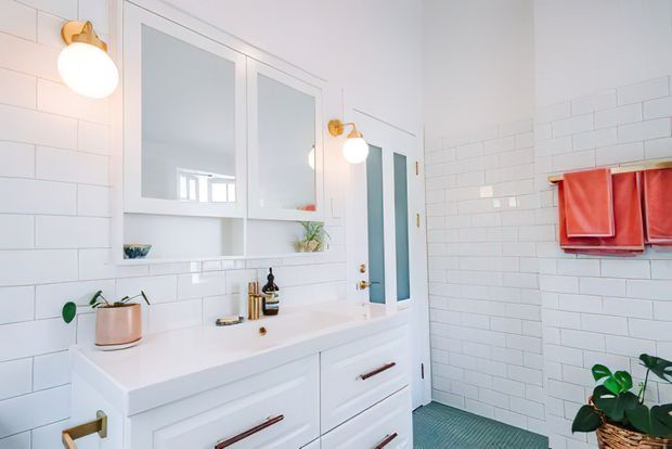 Bright contemporary bathroom with white subway-tile walls, a large white double vanity with integrated basins and wood-toned pulls, mirrored recessed cabinetry, warm wall sconces, and a glass shower enclosure. A textured blue-green floor and terracotta-red towels provide colour accents, with potted plants adding a natural touch.