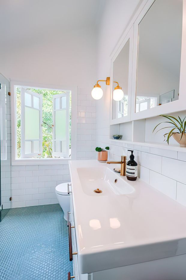 Bright, compact bathroom with white subway-tiled walls, a glossy white integrated vanity and basin, large mirrored cabinetry, brass wall sconces and tapware, and a blue-green mosaic tile floor. An open white-framed window brings in greenery and daylight, while a toilet is partially visible beside the vanity.