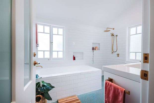 Bright white bathroom featuring two separate bathing areas: a built-in tiled bathtub beneath a window and a freestanding-style white tub in the foreground. White horizontal wall panelling, white subway-tiled bath surrounds, brass shower and tap fixtures, a small blue-green mosaic tile floor, and a coral towel create a clean coastal feel.