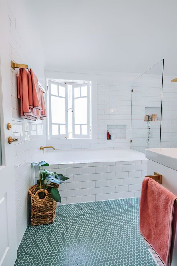 Bright, compact bathroom with white subway-tiled walls and bathtub surround, a white vanity, frameless glass shower screen, and a white-framed window. The floor is finished in small teal-blue mosaic tiles, with brushed brass/gold fixtures and warm coral-pink towels. A woven basket planter adds a natural accent.
