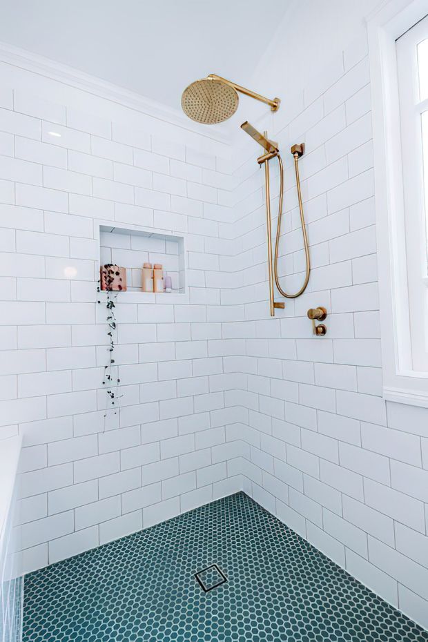 Bright, compact walk-in shower with white subway-tile walls, a dark teal small-format mosaic tile floor, recessed toiletries niche, gold-toned rainfall and handheld shower fittings, and a white-framed window.