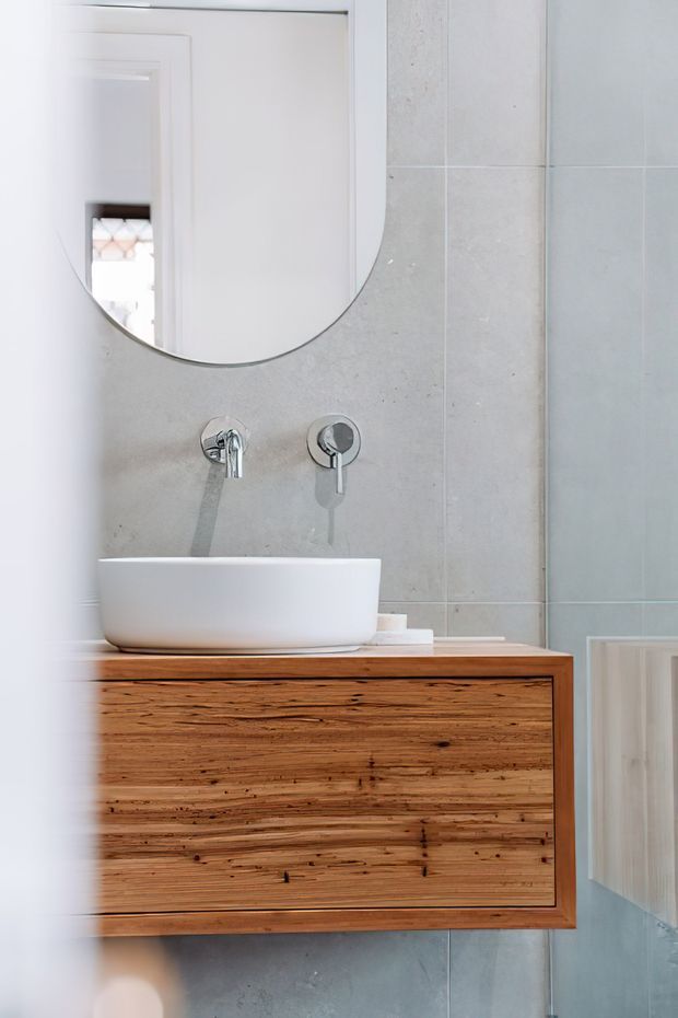 A compact contemporary bathroom vanity with a white vessel basin on a floating timber-veneer cabinet. The wall behind features pale grey tiled surfaces, a wall-mounted chrome tap set, and a rounded rectangular mirror. A white door and trim are partially reflected and visible at the edges.