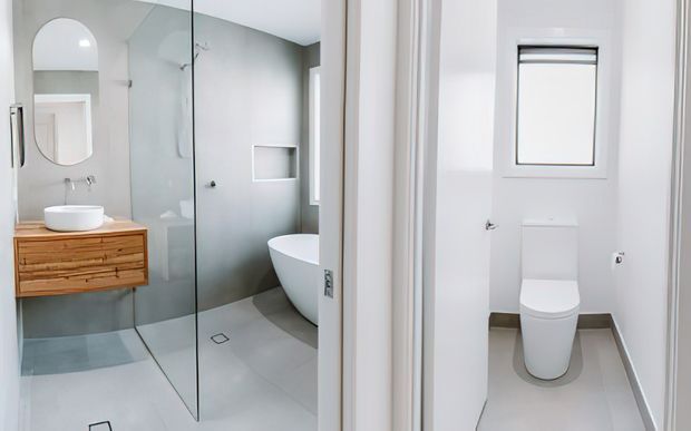 Bright contemporary bathroom suite viewed from the doorway, featuring a walk-in glass shower, freestanding white bathtub, wall-hung timber vanity with vessel basin, rounded-edge mirror, and a separate toilet compartment visible on the right. The space uses pale grey tiled walls and floor with white sanitaryware and clean-lined detailing.
