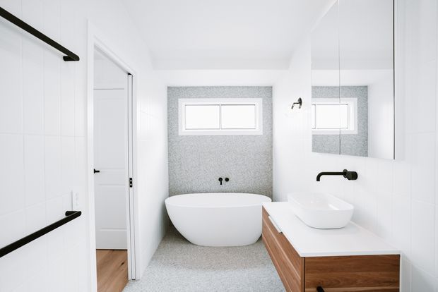 Bright, minimalist bathroom with a freestanding white bathtub set against a speckled grey feature wall, a wall-mounted white basin on a timber vanity, large mirror, white wall panelling, black tapware and hardware, and a small horizontal window. A doorway and timber flooring are visible to the side.