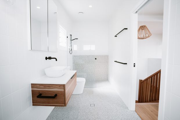 Bright contemporary bathroom with a floating timber-veneer vanity, white vessel basin, wall-hung toilet, frameless glass shower area, and pale terrazzo-like floor and wall surfaces. Black tapware and shower fittings provide contrast. An open doorway reveals a timber staircase and a pendant light beyond.