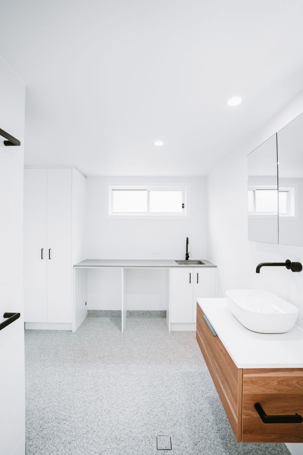 Bright, minimalist white bathroom with a long built-in utility vanity and sink along the rear wall, a freestanding white vessel basin on a warm timber vanity, mirrored wall cabinetry, black tapware and handles, and a light speckled floor finish. The compact room has a high white ceiling, recessed downlights, and a small horizontal window.