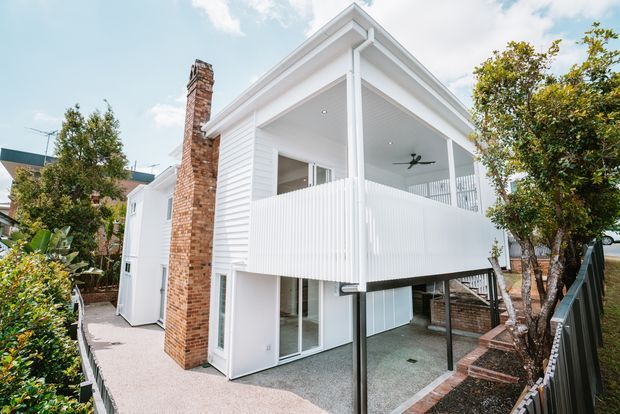 Exterior view of a narrow two-storey residential extension or townhouse with white horizontal weatherboard cladding, a covered upper-level balcony, white vertical privacy balustrade, exposed dark support posts, and a prominent red-brown brick chimney. The lower level has glazed doors and a covered outdoor area, with landscaped planting and fencing around the property.