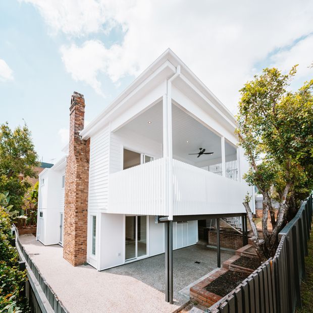 Exterior view of a raised white weatherboard house with a large covered upper-level terrace, vertical privacy balustrade, exposed support posts, brick chimney, lower-level glazing, and a partially enclosed undercroft/carport-like area. The home sits on a compact landscaped site with steps and fencing.