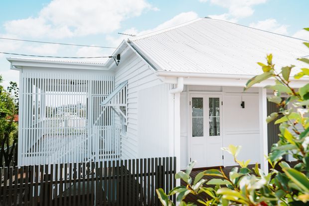 Bright exterior view of a white weatherboard house with a pitched corrugated metal roof, white-framed entry doors, and a covered porch or screened verandah enclosed by vertical slatted screening. Dark vertical fencing and foreground foliage frame the scene.