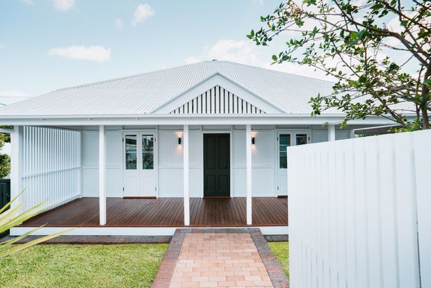 Front exterior of a single-storey white weatherboard-style home with a broad corrugated pitched roof, covered veranda, decorative gable screening, dark entry door, and timber decking. A brick-paved path leads through the front lawn to the veranda, with a white boundary fence partially screening the right side.