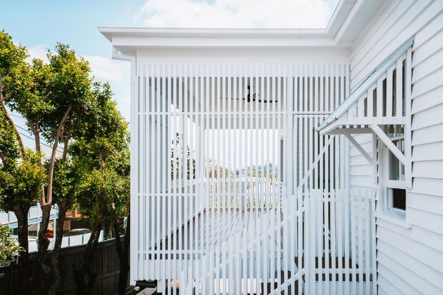 Bright exterior view of a white weatherboard-clad building with a projecting flat-roofed section and a screened outdoor stair or walkway enclosed by closely spaced vertical white battens. A small awning shelters a window, with landscaped trees visible at the side.