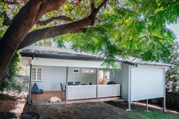 Single-storey light-grey weatherboard house viewed from the garden, with a covered timber front or rear deck, white vertical-slat balustrade, enclosed porch, tiled pitched roof, and an elevated enclosed side structure. Mature trees surround the home.