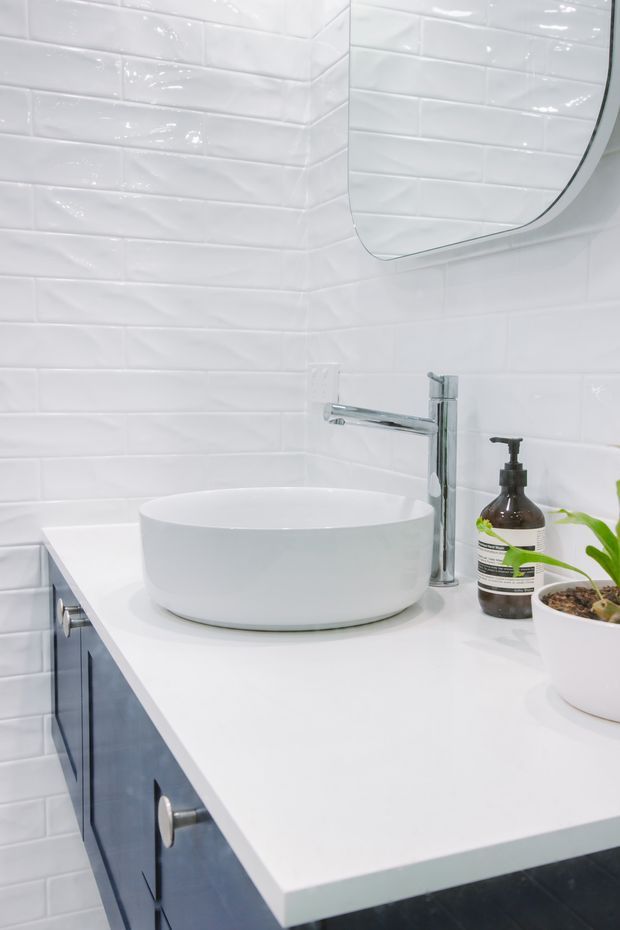 Close-up of a bright contemporary bathroom vanity featuring a white vessel basin on a white benchtop, chrome wall-mounted tapware, dark blue fixed cabinetry, glossy white subway wall tiles, and a rounded-edge mirror. A soap dispenser and small potted plant sit on the vanity.