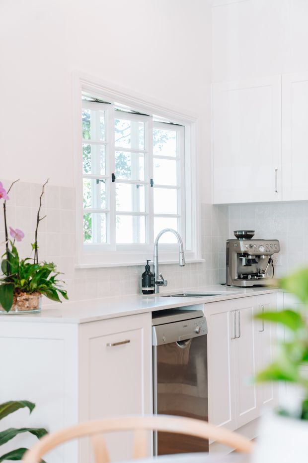 Bright white kitchen with shaker-style cabinetry, pale tiled splashback, white benchtops, stainless-steel dishwasher, undermount sink and curved chrome tap beneath a multi-pane window. A coffee machine and potted orchids add detail, with soft natural light creating an airy, fresh atmosphere.