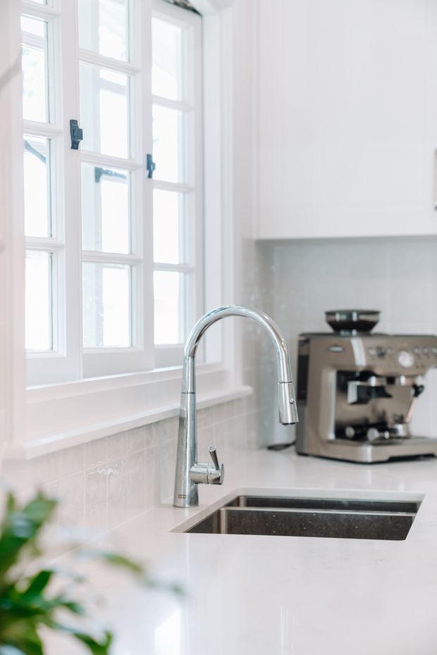 Bright, light-filled kitchen close-up featuring a polished chrome gooseneck faucet over a double-basin undermount sink, white cabinetry, pale tiled splashback, and large white-framed multi-pane windows. A stainless steel espresso machine sits on the rear counter, with soft greenery partially visible in the foreground.