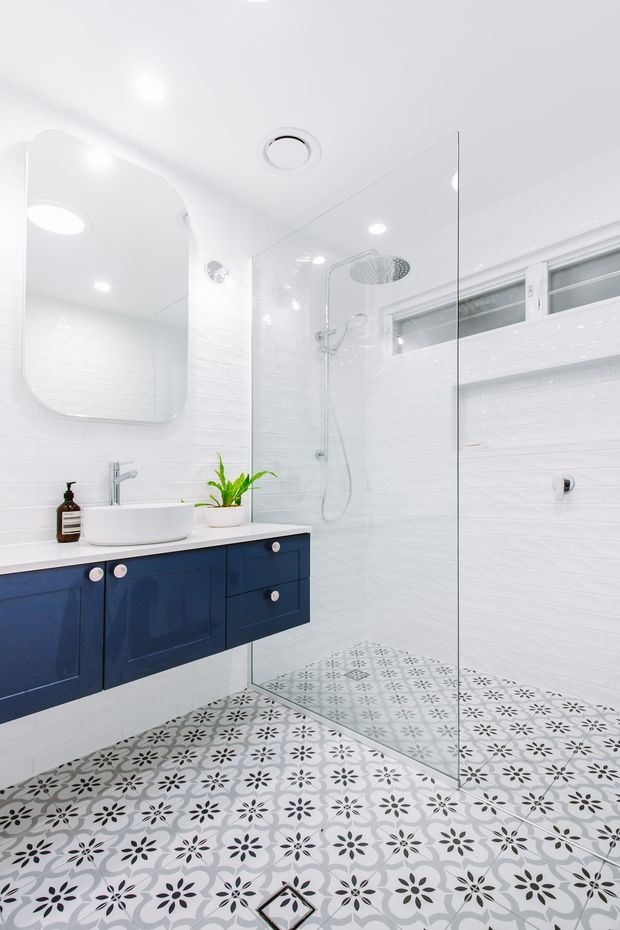 Bright contemporary bathroom featuring a floating navy-blue vanity with a white vessel basin, large rounded-edge mirror, white tiled walls, a frameless glass walk-in shower, and patterned encaustic-style floor tiles in white, grey, and black. Recessed ceiling lighting and a horizontal window create an airy, clean atmosphere.