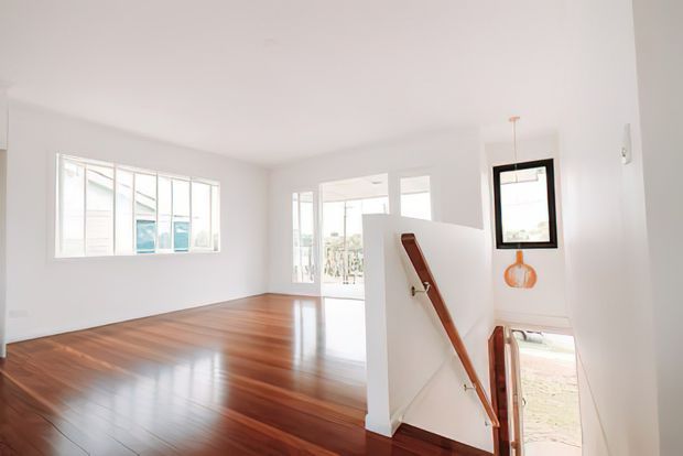 Bright, minimalist open-plan interior viewed from a stair landing, featuring polished dark timber flooring, white walls and ceiling, large glazed windows and doors, and a simple timber handrail descending to a lower level. The space appears airy and uncluttered with abundant natural light.
