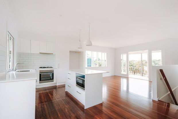 Bright open-plan kitchen and living area with white cabinetry, a central island, white subway-tile splashback, stainless-steel oven and cooktop, polished timber flooring, pendant lights, and extensive glazed doors and windows. A staircase is visible at the right, with a balcony or exterior view beyond the living area.