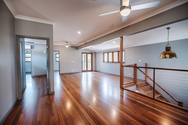 A bright, open-plan upper-level interior with polished timber flooring, grey walls, white ceiling trim, recessed lighting, and a ceiling fan. A timber-and-metal balustrade overlooks a lower level, while glazed timber doors and windows bring in natural light. A copper pendant light adds a warm accent near the stair void.