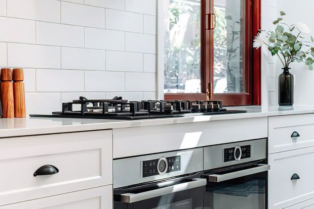 Bright, compact kitchen focused on a white tiled splashback, white cabinetry with dark cup pulls, a pale benchtop, black gas cooktop, and integrated stainless-steel ovens. A dark timber-framed window and a simple vase provide contrast.