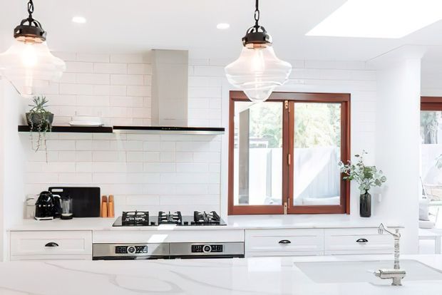 Bright, airy contemporary white kitchen with white subway-tile walls, white cabinetry, black floating shelves, stainless-steel cooking appliances, and a white stone island benchtop. Timber-framed windows and glass pendant lights add warmth and contrast, while a skylight brings in additional natural light.