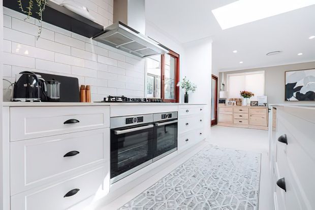 Bright, spacious white kitchen with shaker-style cabinetry, black hardware, a stainless-steel rangehood, and a wide double oven beneath a gas cooktop. White subway-tile splashback, dark benchtops, timber-framed glazing, skylights, and a patterned runner define the room. A furnished adjoining living or dining area is visible beyond the kitchen.