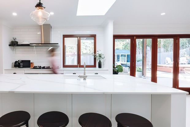 Bright, contemporary white kitchen with a large white island and dark bar stools. The room features white horizontal wall panelling, white cabinetry, a dark countertop or shelf detail, timber-framed windows and glazed folding doors opening to an outdoor area. A skylight and pendant light provide additional natural and decorative illumination.