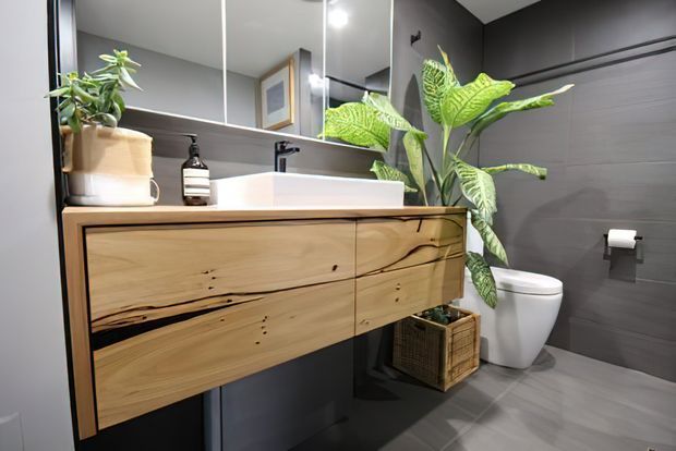 Contemporary bathroom featuring a wall-mounted natural timber vanity with integrated white basin, large mirrored cabinetry, dark grey wall and floor tiles, a white toilet, and tropical indoor plants.