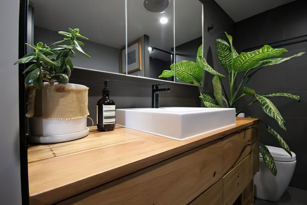 A compact, stylish bathroom vanity with a white vessel basin and matte black tap set on a warm timber drawer vanity. Dark charcoal walls, a large mirror, integrated lighting, and leafy indoor plants create a moody yet organic atmosphere.