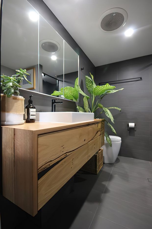 A contemporary bathroom featuring a floating natural timber vanity with integrated drawers, a white vessel basin, matte black tapware and fixtures, large mirrored cabinetry or wall panels, and dark grey tiled walls and floor. Indoor plants add a natural accent, while recessed lighting and a ceiling ventilation grille provide bright task lighting and ventilation.
