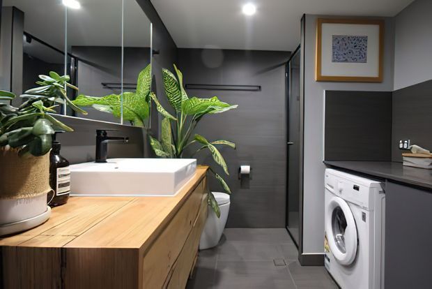 A contemporary combined bathroom and laundry room. The foreground features a timber vanity with a white vessel basin and matte black tapware, beside a large wall mirror and indoor plants. At the rear is a dark tiled shower enclosure, while the right side contains white front-loading laundry appliances beneath dark cabinetry and shelving. Recessed ceiling lights and charcoal finishes create a refined, moody interior.