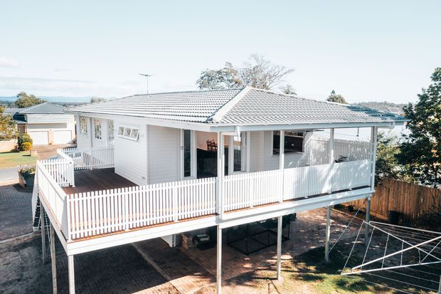 Elevated single-storey suburban home with a light weatherboard exterior, pale grey tiled hipped roof, and a large wraparound white picket-balustraded deck. The house is raised on slender posts, with a covered outdoor area beneath and a residential neighbourhood visible in the background.