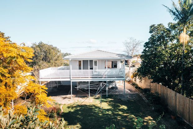 Rear exterior view of a raised single-storey home with a light corrugated hipped roof, pale exterior walls, and a broad covered veranda with white balustrades. The house sits above an open undercroft and overlooks a fenced garden with established trees and landscaping.