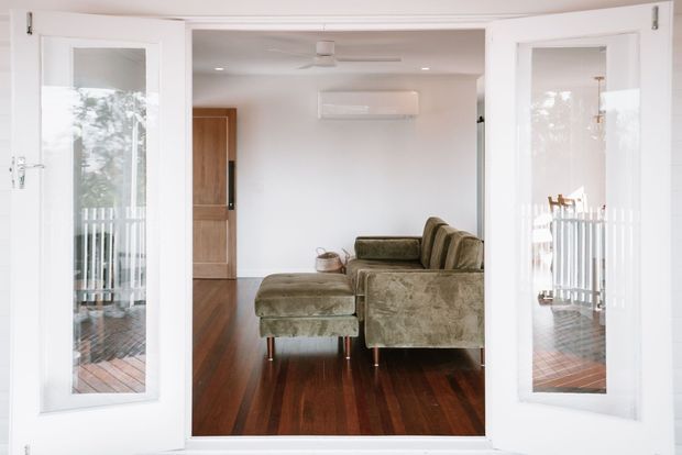 View through open white-framed glazed double doors into a compact living room. The room has polished dark timber flooring, white walls and ceiling, a muted olive-green upholstered sofa with matching ottoman, a wall-mounted split air conditioner, ceiling fan, and timber door. Reflections and views through the side glazing show an adjoining timber deck and white balustrade.