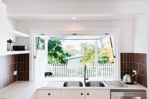 Bright compact kitchen with white cabinetry and benchtops, a double stainless-steel sink beneath a large open casement window, and dark brown tiled splashback wrapping the walls. Built-in shelving and a kettle are visible, with a leafy residential outlook beyond the window.