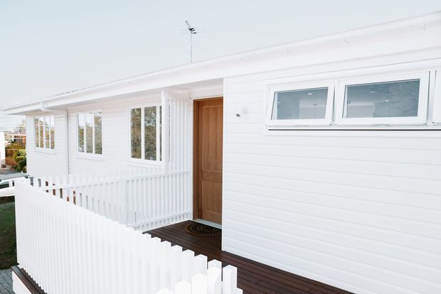 Exterior view of a bright white single-storey house with horizontal weatherboard cladding, a low-pitched roof, timber entry door, white-framed windows, and a dark timber deck and entry walkway enclosed by white picket-style balustrades.