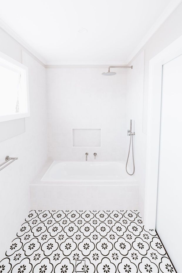 Bright, compact white bathroom with a built-in bathtub and wall-mounted shower fixtures. The walls, ceiling, bath surround and window trim are white, while the floor features distinctive black-and-white floral patterned tiles. Chrome fixtures and a recessed wall niche add functional detail.