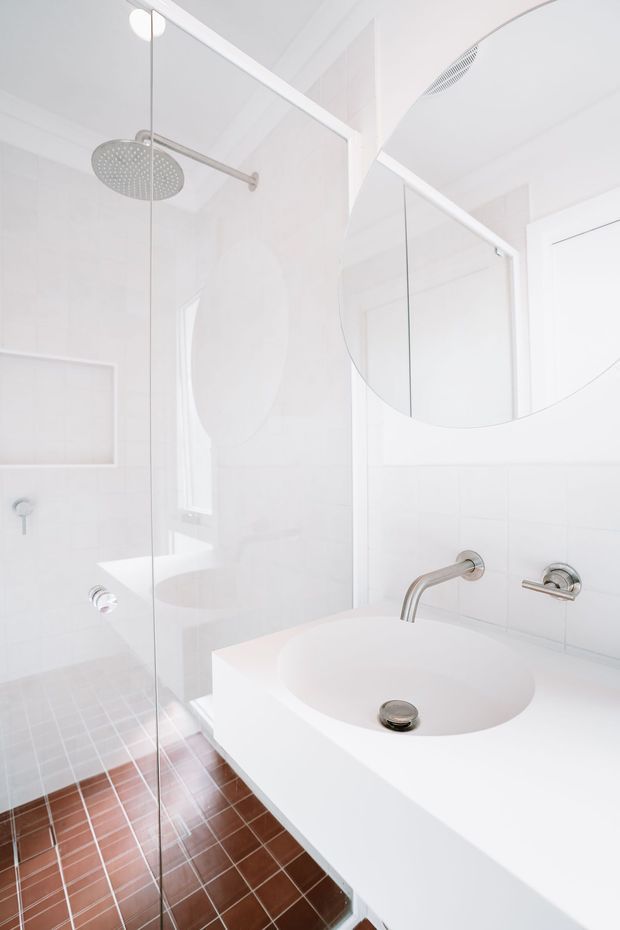 Bright, minimalist bathroom with a frameless glass walk-in shower, round rainfall showerhead, wall-mounted brushed metal tapware, a large curved mirror, white tiled walls, and a white integrated vanity basin. Small reddish-brown square floor tiles provide contrast against the predominantly white fit-out.